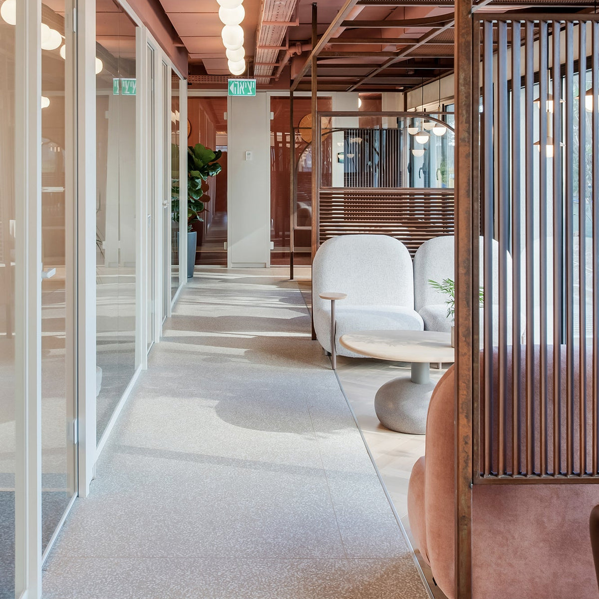 A modern interior hallway with glass-walled offices on the left, pendant lights hanging from a terracotta ceiling, and lounge seating with white curved chairs and a pink upholstered chair on the right.