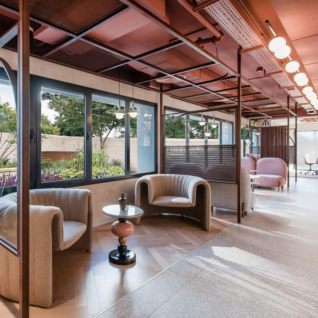 A modern interior lounge area with two beige upholstered armchairs facing a small round table, large black-framed windows, exposed wood ceiling beams, and warm pendant lights.