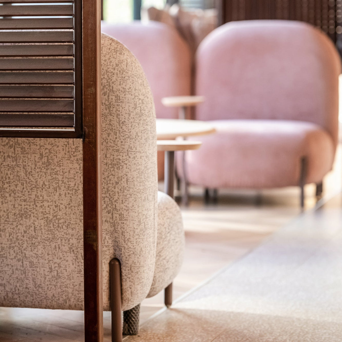 A modern interior scene with upholstered lounge chairs in beige and blush pink, separated by a dark wooden slatted screen, with warm lighting and a softly blurred background.
