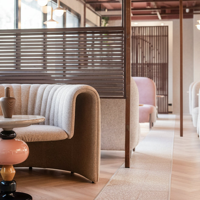 A modern curved upholstered sofa in a warm beige fabric, styled in a contemporary indoor lounge setting with a wooden slat partition, pendant lights, and a decorative pink and black side table in the foreground.