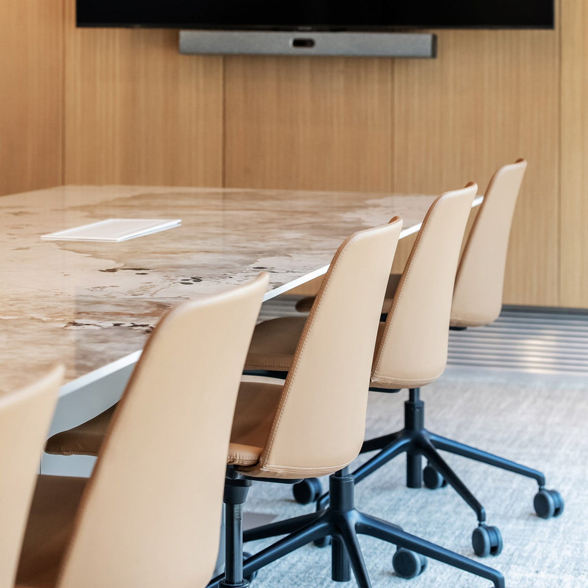 A modern conference room setup featuring a large black wall-mounted screen above a marble-top meeting table with beige swivel chairs.