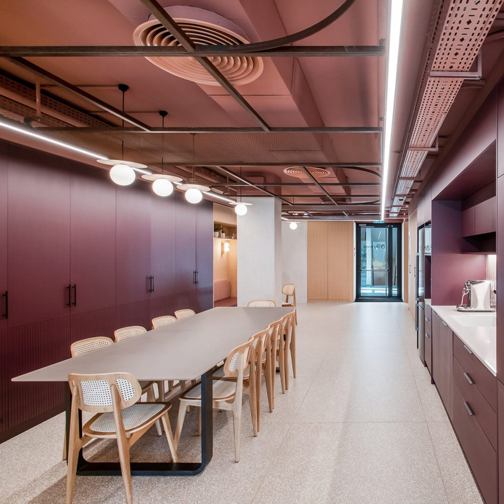 A modern interior space with a long rectangular dining table and chairs on the left, and a kitchenette with dark plum cabinets and light wood counters on the right. The room has a high ceiling with exposed industrial ductwork and linear lighting, and a light stone floor.