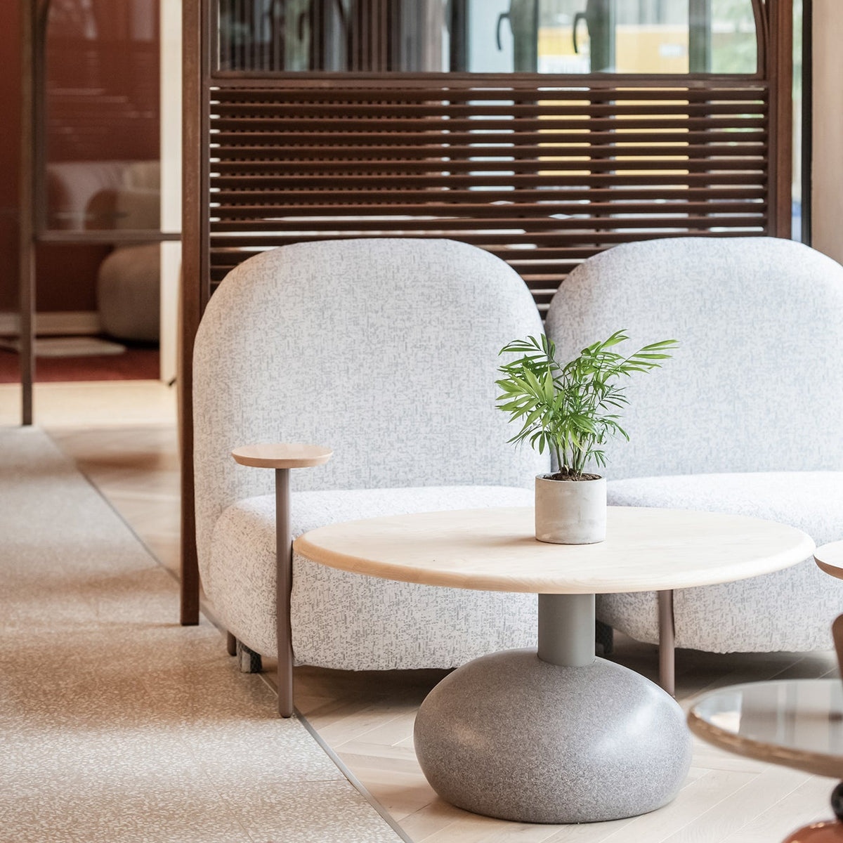 A modern lounge area featuring a light gray upholstered two-seat sofa, a round light wood coffee table with a gray pedestal base, and a small potted green plant, set within a contemporary interior with warm wood framing and glass partitions.