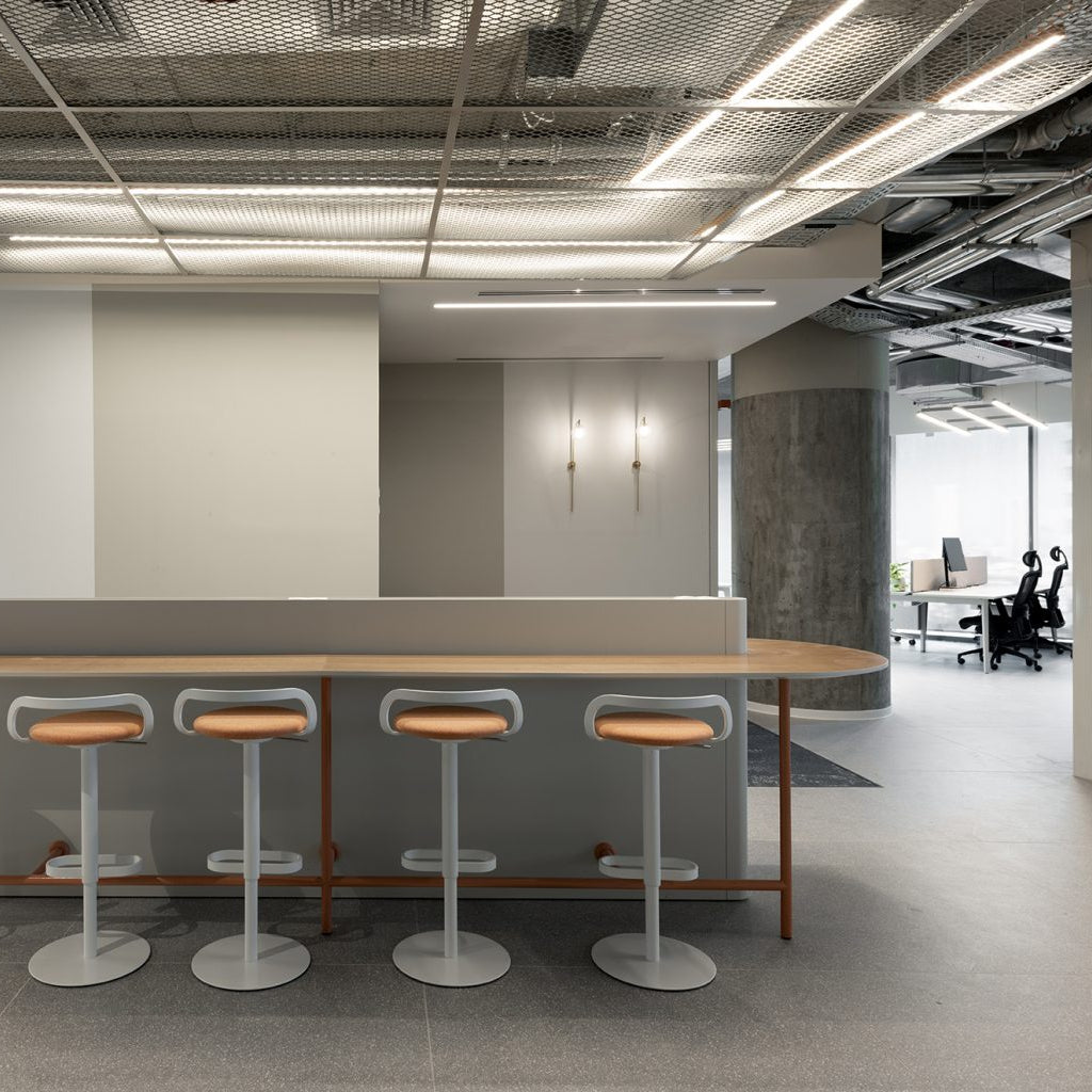 A modern industrial-style office interior featuring a long bar-height counter with five bar stools, set against a minimalist concrete and glass office space.