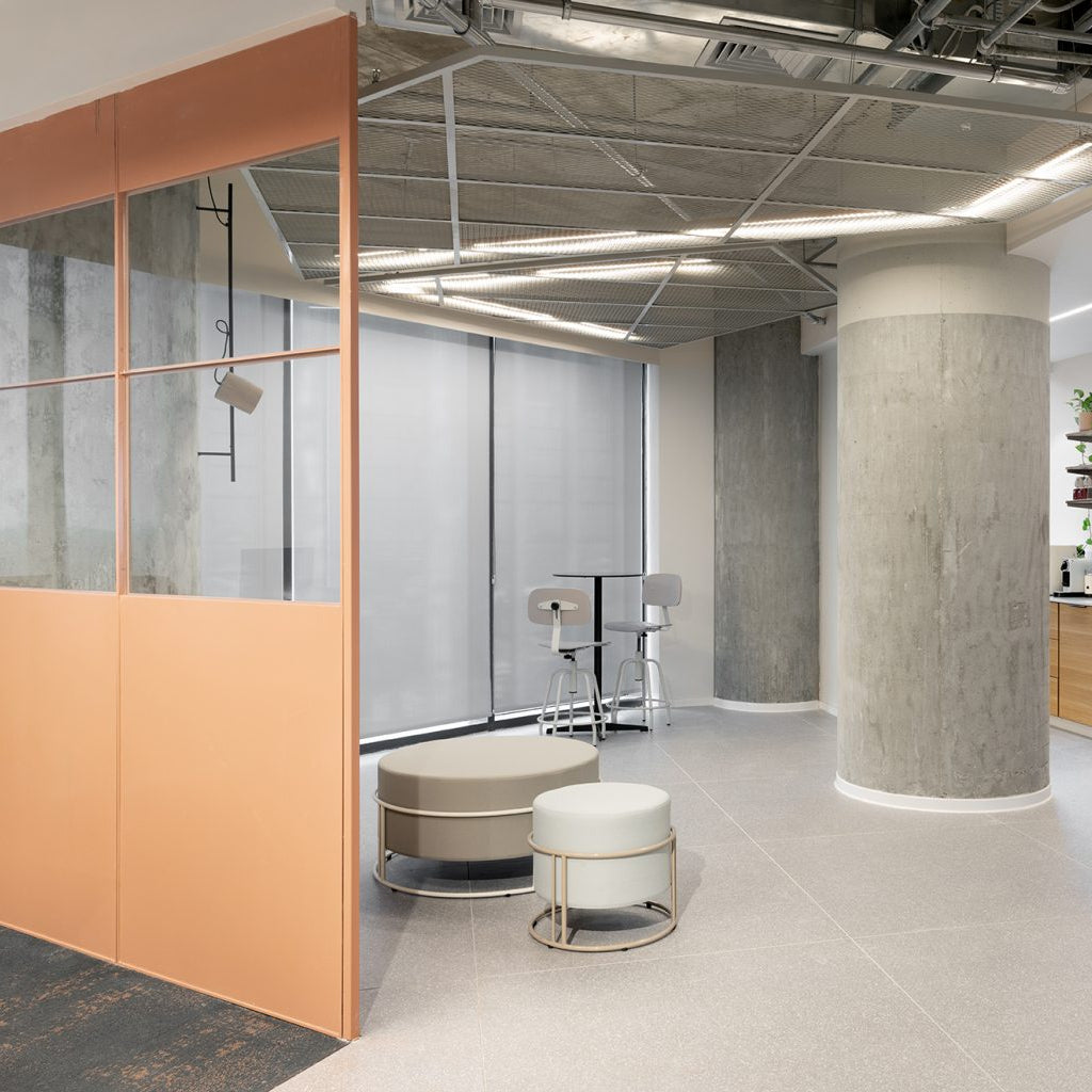 A modern office interior with a peach-colored glass partition wall, a large concrete column, light gray flooring, and an open-plan workspace with stools and a kitchen area in the background.