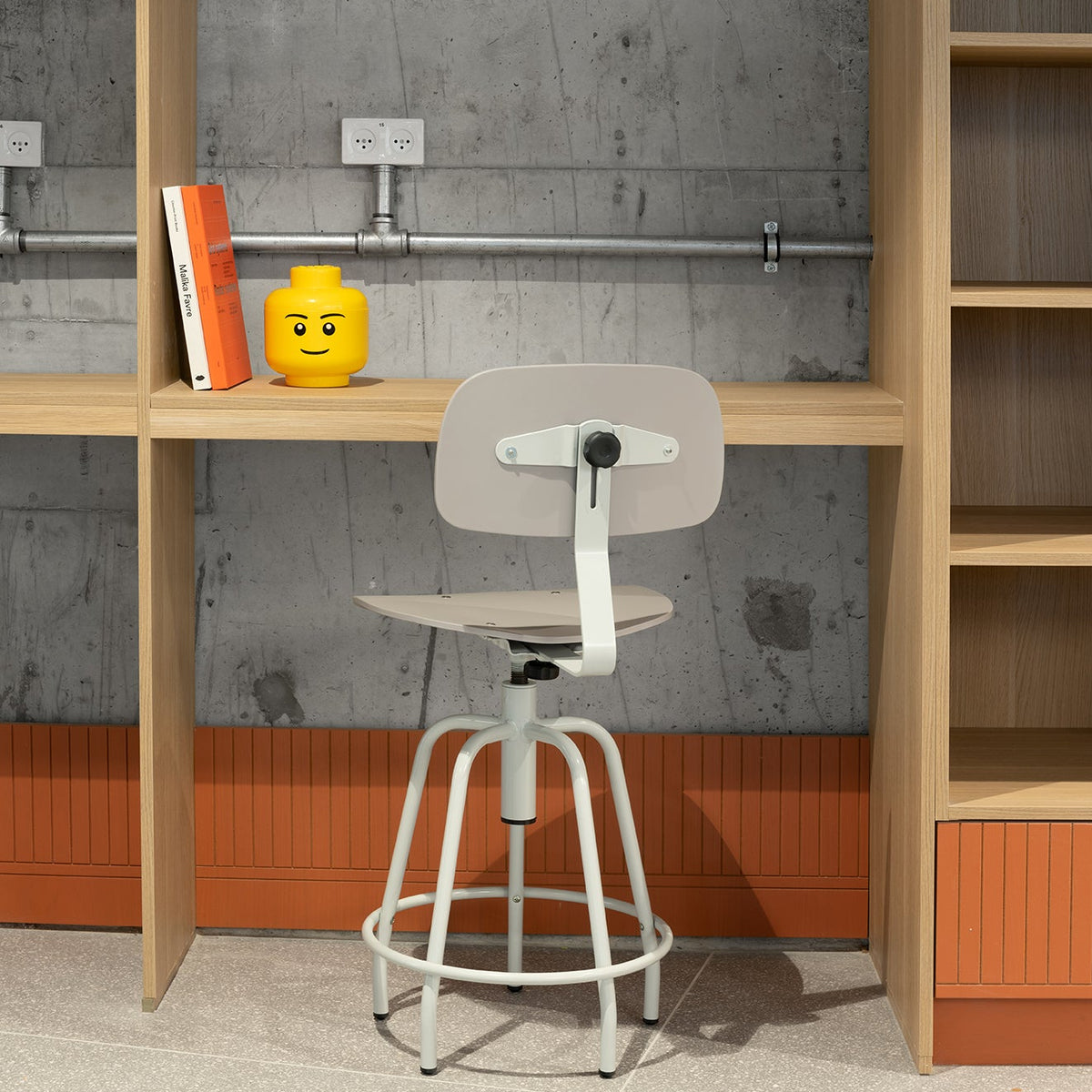 A modern white swivel stool with a backrest, positioned in front of a light wood desk and shelving unit against a textured gray concrete wall.