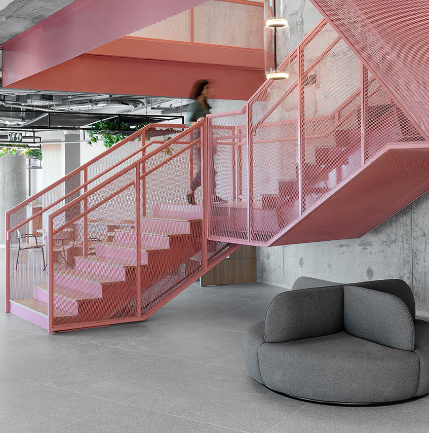 A modern interior scene with a pink metal staircase and matching pink ceiling structure in a concrete-walled office space. A person is walking up the stairs, and a round gray lounge chair sits in the foreground.
