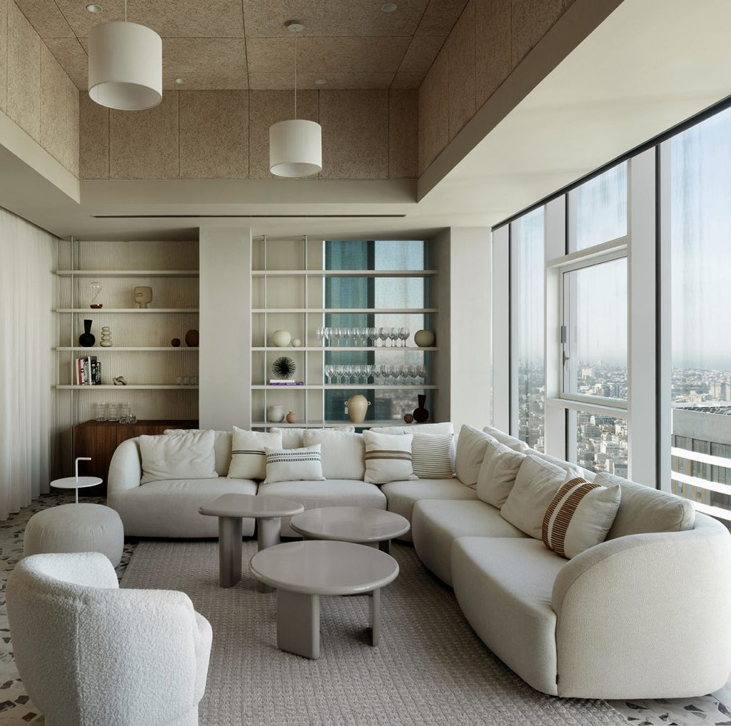 A modern living room featuring a curved cream sectional sofa, matching armchair, round coffee tables, built-in shelving, and large floor-to-ceiling windows overlooking a city skyline.