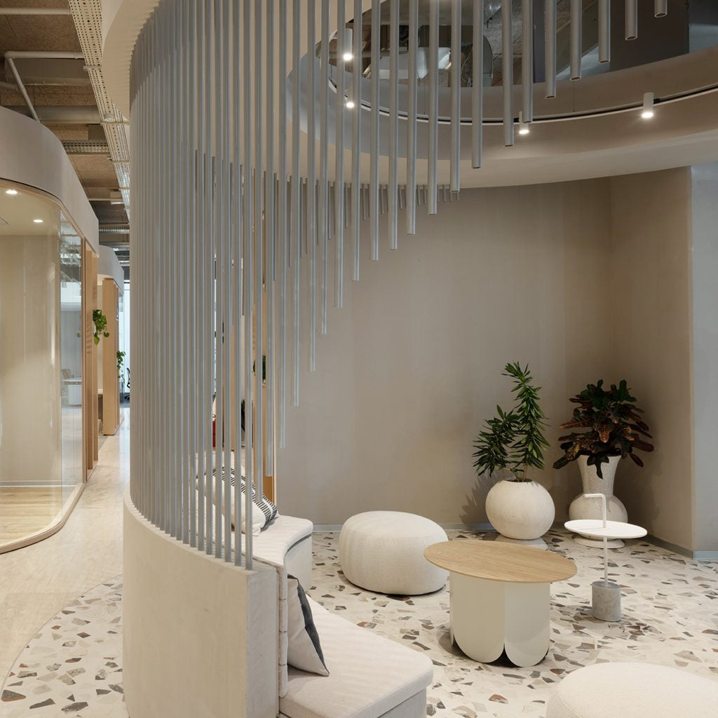 A modern interior space with a curved glass-walled meeting room on the left and a seating area on the right featuring a curved white sofa, round white poufs, small white tables, and a decorative vertical slat screen divider. The room has light wood flooring, beige walls, and potted plants.