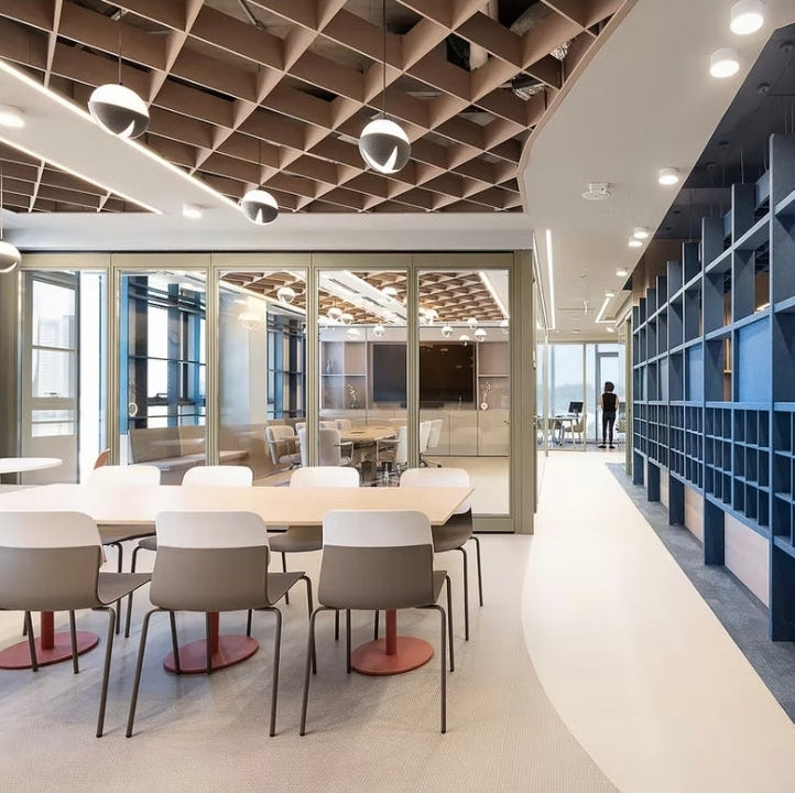 A modern office interior with a long blue shelving unit along the right side, white dining tables and chairs in the foreground, glass walls, and a geometric wood ceiling.