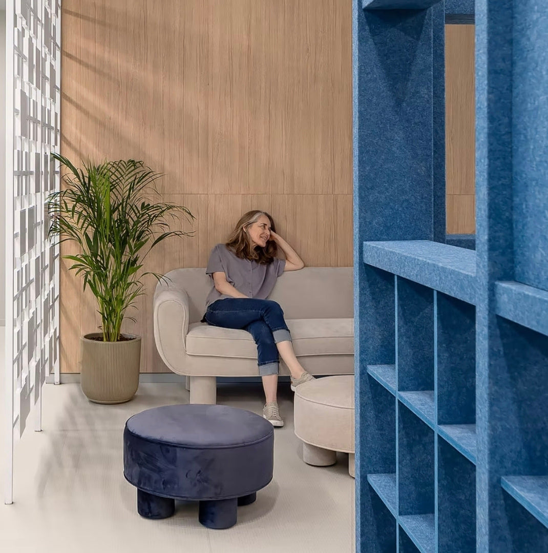 A woman sits on a light beige sofa in a modern interior with blue shelving, a wood-paneled wall, pendant lights, and a potted plant.