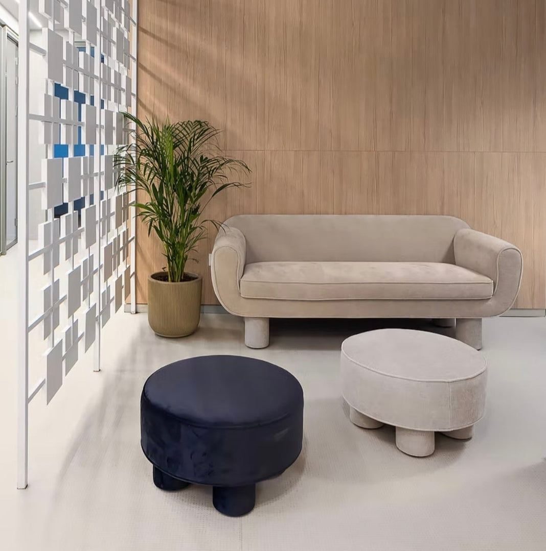 A modern interior lounge area featuring a beige upholstered sofa, two round ottomans (one dark navy velvet, one light beige fabric), a potted green plant, a white geometric screen wall, and a dark blue ceiling with exposed pipes and pendant lights.