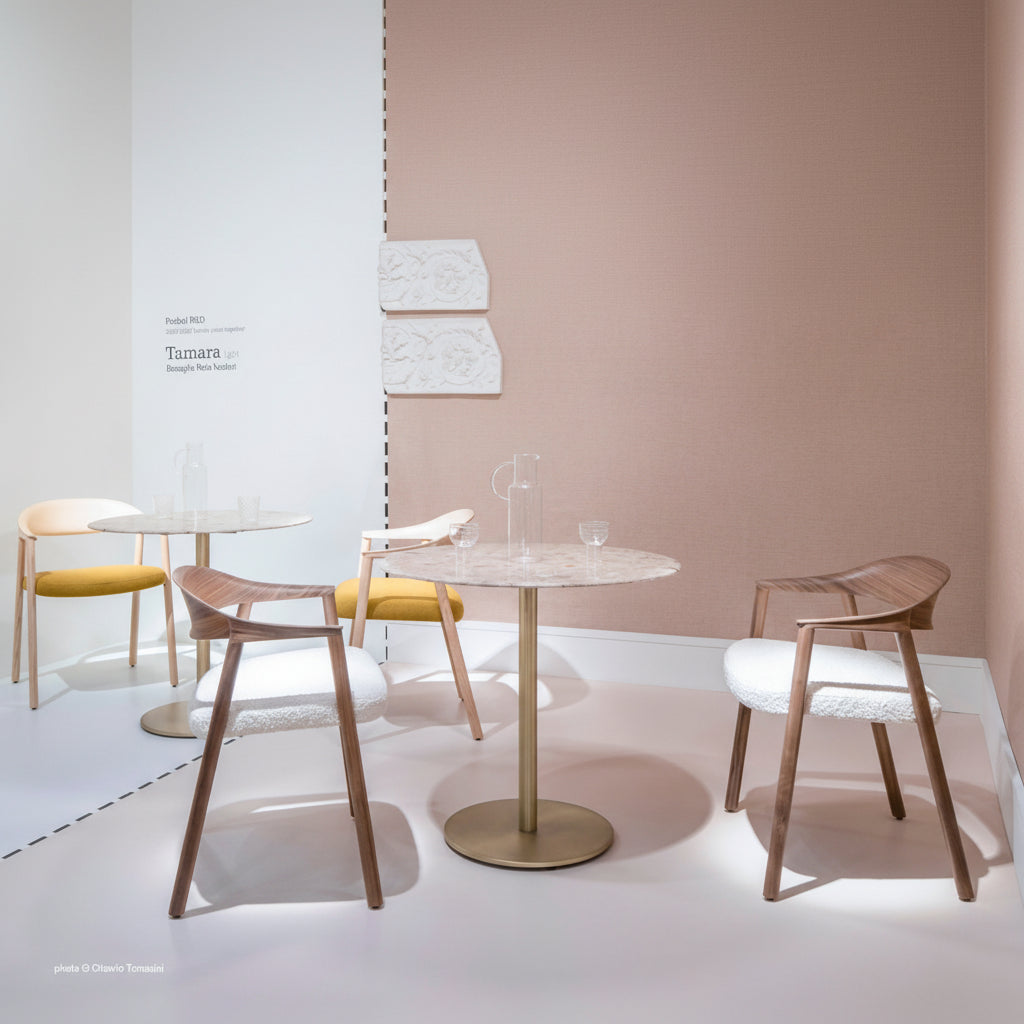 A modern dining set featuring a round marble-top table with a brushed brass pedestal base, surrounded by two wooden armchairs with white textured upholstery and two chairs with yellow seat cushions, set against a warm beige wall.