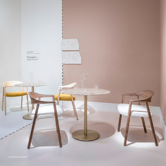 A modern dining set featuring a round marble-top table with a brushed brass pedestal base, surrounded by two wooden armchairs with white textured upholstery and two chairs with yellow seat cushions, set against a warm beige wall.